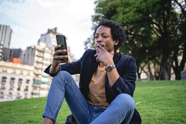 Carefree hipster smoking a cigarette at a park while looking at his mobile phone