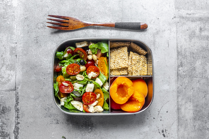 Lunch box with rocket salad with colored tomatoes, mozzarella and nuts, crispbread and apricots, wooden fork on concrete surface