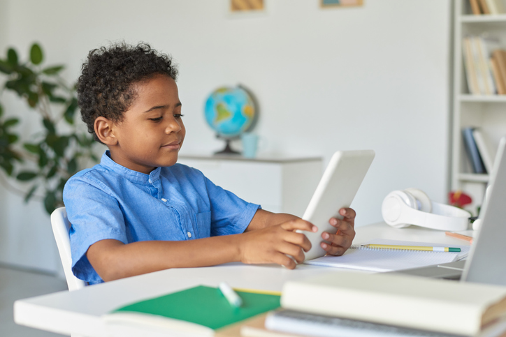 Modern African American schoolboy sitting at desk and using tablet for education at home