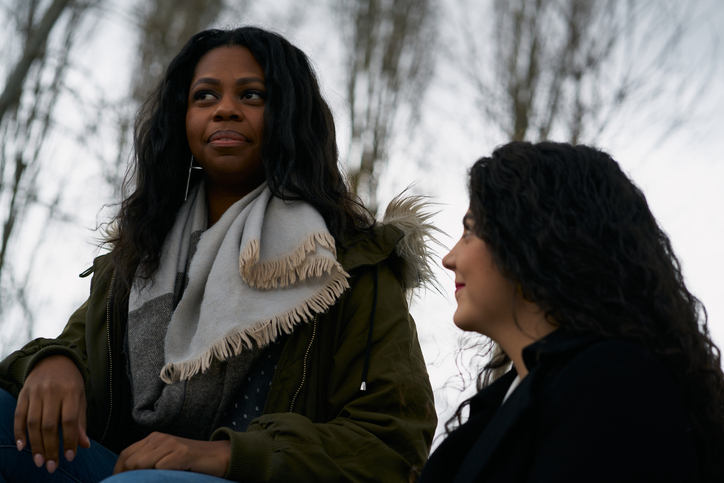 Pair of female friends wearing winter coats and talking sitting on a red bench in a park. Friendship concept