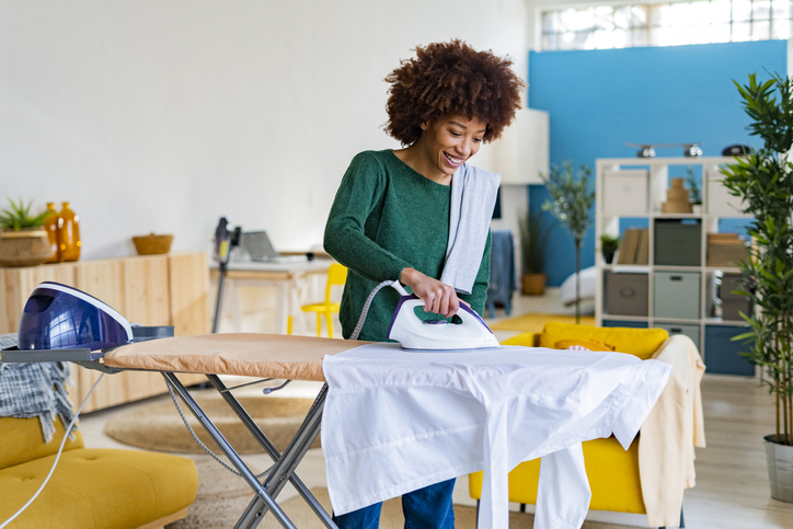 Smiling young Afro woman ironing shirt on board in living room