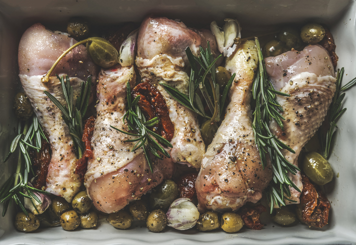 Raw marinated chicken drumsticks in baking sheet with olives, capers, dried tomatoes and rosemary