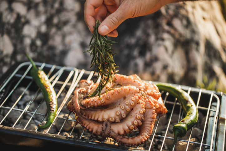 Close up of manâs hand with rosemaryâs bunch cooking octopus on barbecue.