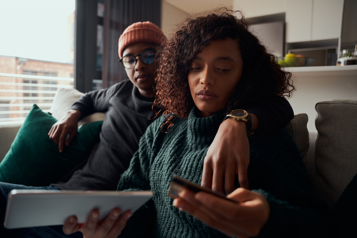 Adult couple, multi-ethnic, concerned about making online payment on tablet sitting sofa in modern apartment