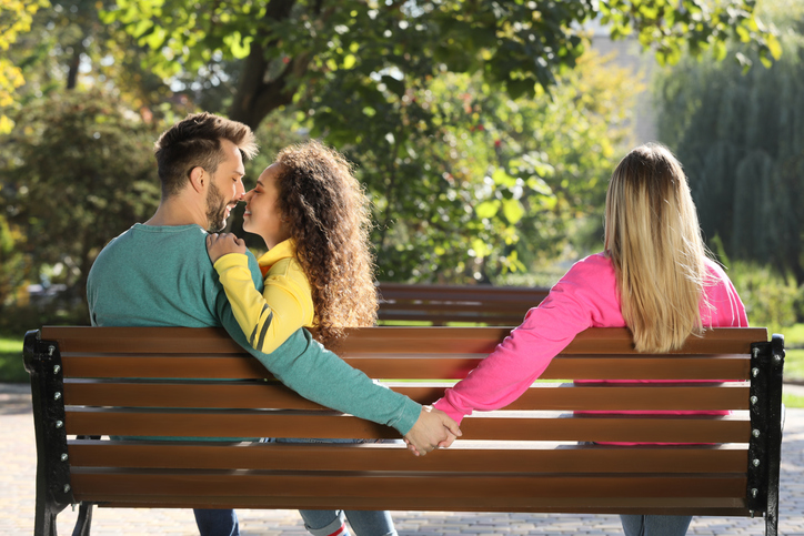 Man holding hands with another woman behind his girlfriend's back on bench in park. Love triangle involved in mate poaching