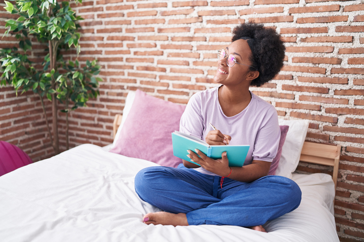 African american woman writing on notebook sitting on bed at bedroom with zen energy