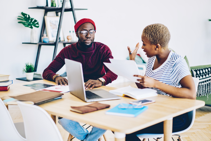Serious dark skinned male and female colleagues having discussion during working process disappointed with planning, angry african american woman quarrel with hipster guy checking documents at desktop
