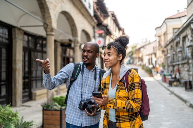 Young african couple of tourists with smartphone looking for destination and taking pictures on camera while walking on the street in an old town in Europe