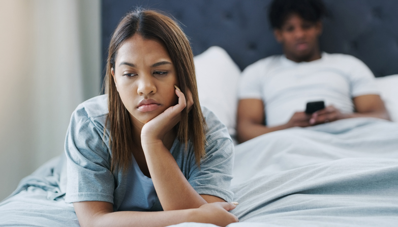 Shot of a woman looking upset while lying on her bed with her boyfriend using his cellphone in the background