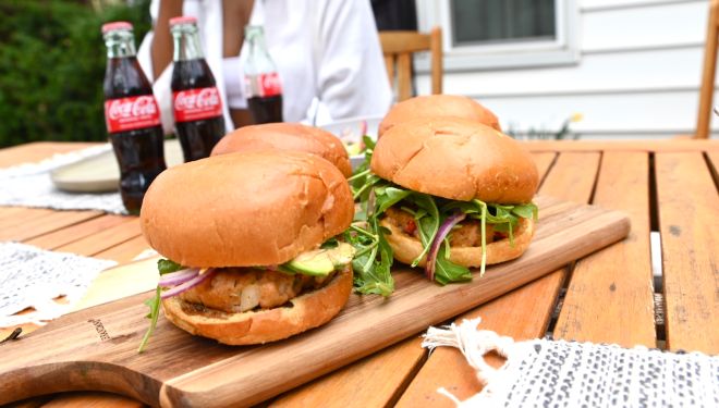 Chef Scherise's jerk salmon burgers, the finished product. They are dressed with fresh toppings and sriracha aioli. Three bottles of Coca Cola sit behind the jerk salmon burgers.