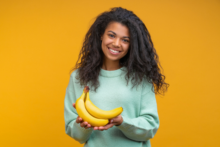 Studio shot of happy smiling attractive young woman posing with a bunch of fresh ripe bananas in hands isolated over bright colored orange yellow background