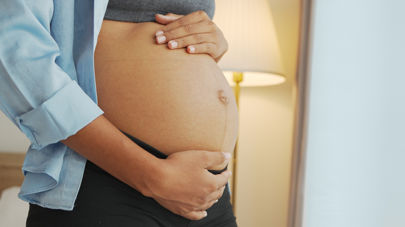 Black African American Pregnant woman holding her belly with both hands in a cozy room, showing care and anticipation.