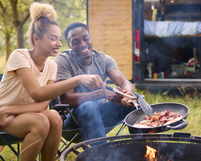 Couple Camping In Countryside With RV Cooking Bacon And Eggs For Breakfast Outdoors On Fire