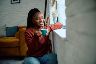 Smiling black woman day dreaming while having a cup of coffee by the window.