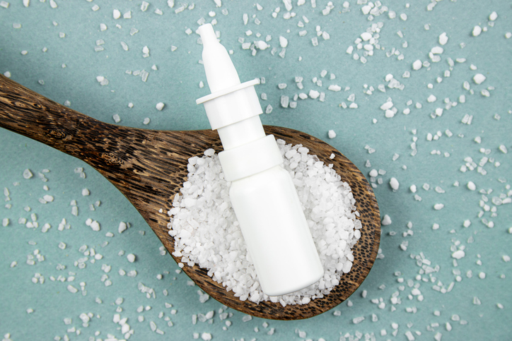 Homemade saline nasal spray bottle on scattered sea salt grains and wood spoon on blue background, studio shot.