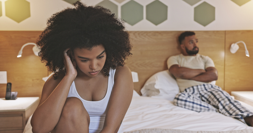 A couple after a fight in their bedroom. Upset young woman sitting on her bed with indifferent angry boyfriend in the background. Pregnancy scare in unhappy unfulfilled relationship. Marriage issues