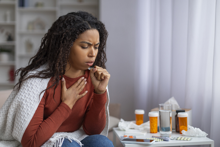 Black woman suffering cold, sitting wrapped in blanket and coughing at home