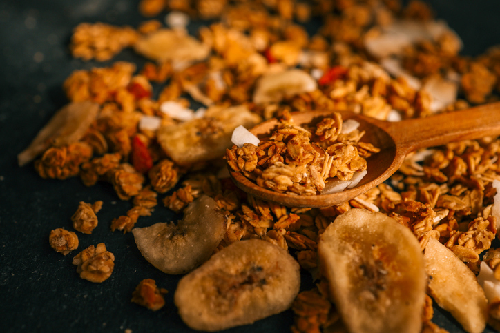Overhead view of a wooden spoon with scattered granola, dried banana chips, and coconut flakes on a dark textured background