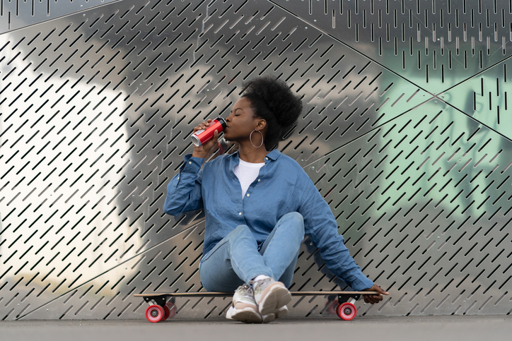 Tired afro american skateboarder girl refreshing after longboarding sit on skate drink soda beverage