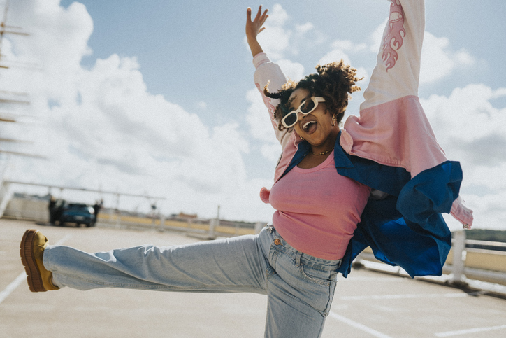 Cheerful curly hair woman in sunglasses and letterman jacket dancing at parking lot under cloudy sky