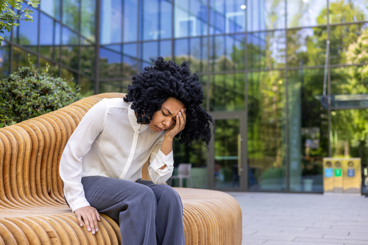 Sad woman sitting on a bench, overwhelmed woman has severe headache, tired and overwhelmed in depression sitting outside office building, holding hand on head