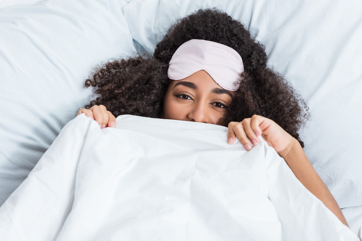 happy young african american woman with blindfold on forehead covering face by blanket and laying in bed during morning time at home