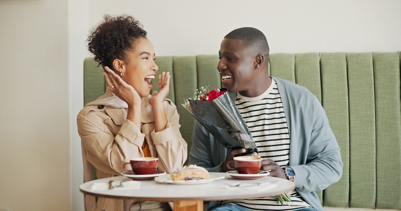 Flowers, excited or couple at coffee shop for surprise on romantic anniversary date together. Wow, roses or shocked interracial people bonding at cafe or restaurant for love announcement at breakfast