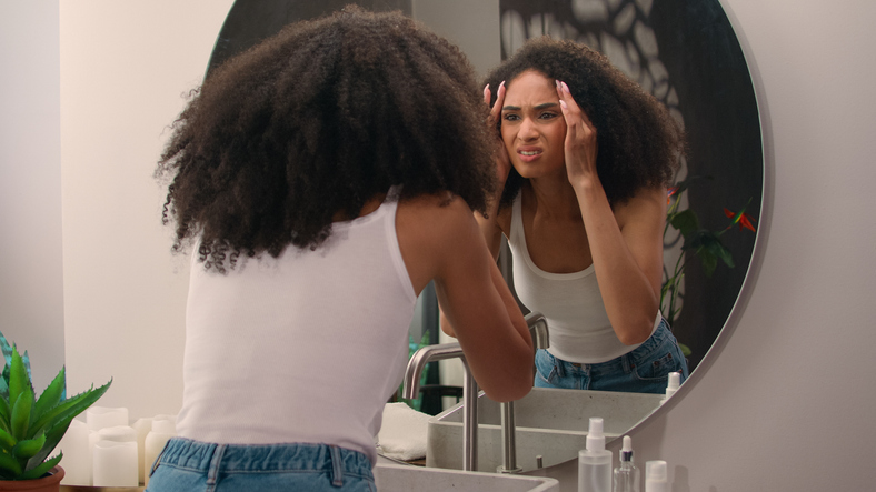 African American beautiful attractive female girl woman come in bathroom home looking at mirror reflection check skin care problem acne wrinkles dermatology trouble upset with skincare facial pimple