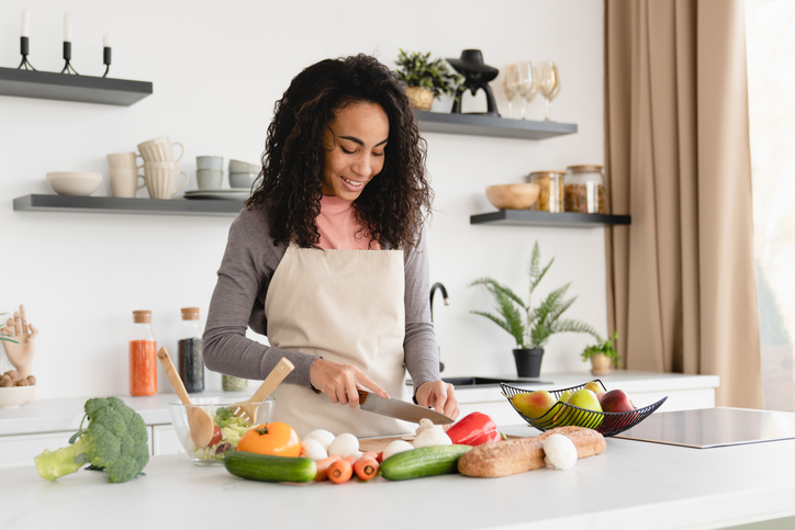 Happy young african chef housewife woman cooking making preparing food dinner lunch at home kitchen, cutting vegetables for salad. Vegan vegetarian meal concept
