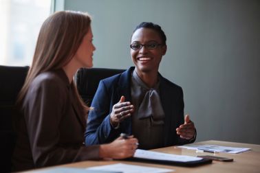 Mid adult Black ethnicity woman businesswoman financial advisor smiling talking plans ideas with paperwork financial planning with mature woman work colleague in businesswear business meeting at board room conference table in office