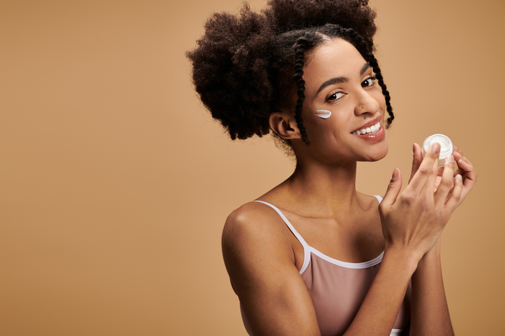 Young woman applies skincare cream with a joyful smile in a warm, minimal setting