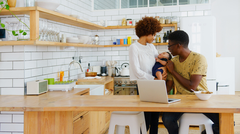 Busy Family In Kitchen At Breakfast With Father Working On Laptop And Mother Caring For Baby Son