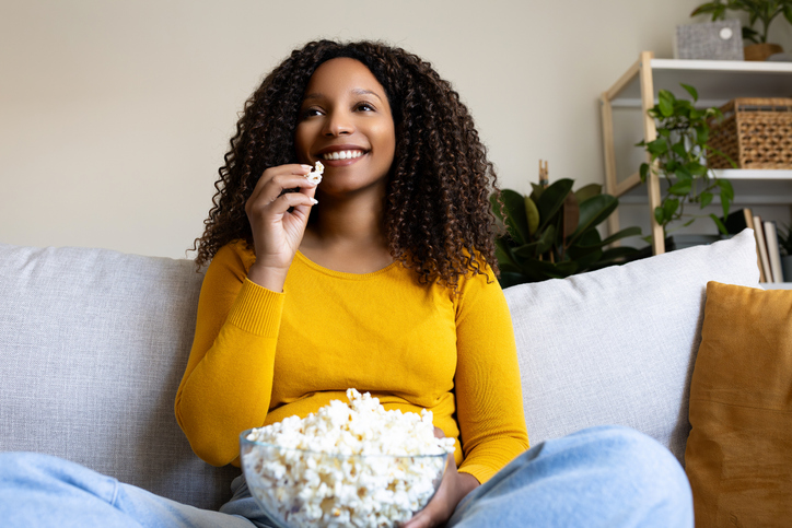 African American woman watching movie at home eating popcorn, sitting on the couch smiling.