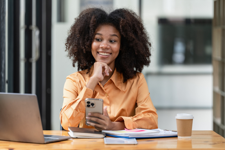 Portrait of a beautiful confident businesswoman using a laptop computer holding a mobile phone sitting in a modern office. Smiling African American freelancer working online from home.