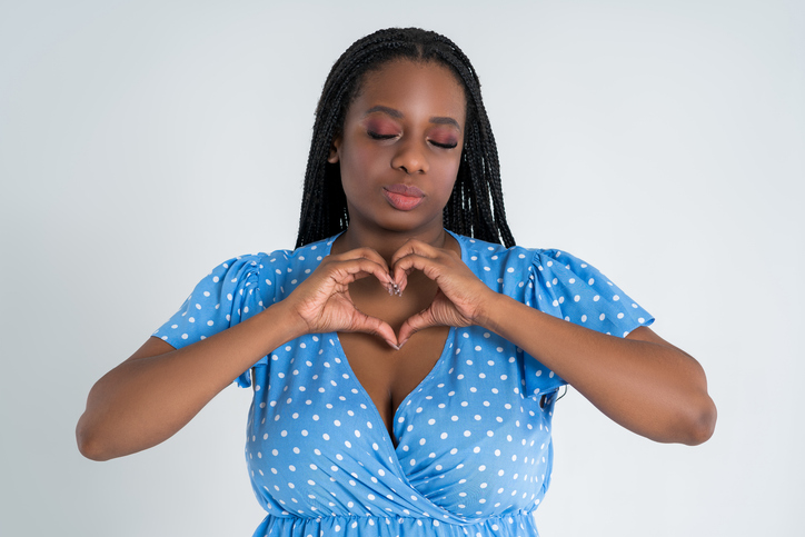 Plus size female model posing in blue dress on white background and showing heart sign, young African woman with curvy figure and pigtailed hairstyle, afro braids