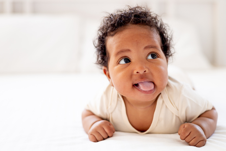 a close-up portrait of a small African-American baby girl in a white bodysuit on the bed at home, a funny six-month-old black newborn baby lies on her tummy and shows her tongue