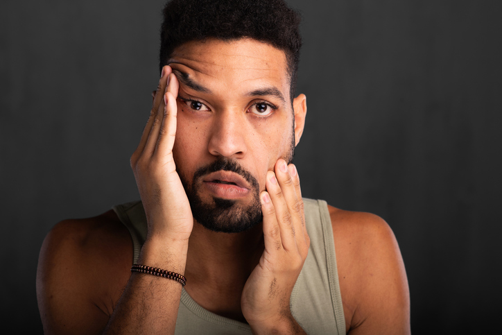 Handsome man stretching his face skin, men’s beauty standards and skincare. Portrait of a young man dissatisfied with his appearance, skin, or beauty.
