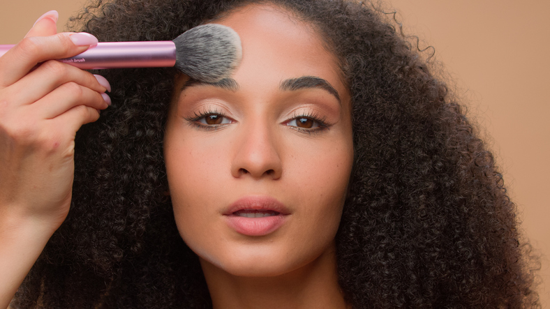 Close up portrait beige studio background beautiful African American woman with perfect smooth skin apply powder looking at camera attractive ethnic girl powdering face make-up cosmetics blush beauty