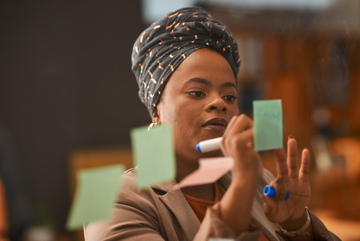 Black woman, writing and sticky note for schedule, planning and brainstorming, creative and strategy on glass board. African person, employee and reminder, tasks and agenda for startup in office