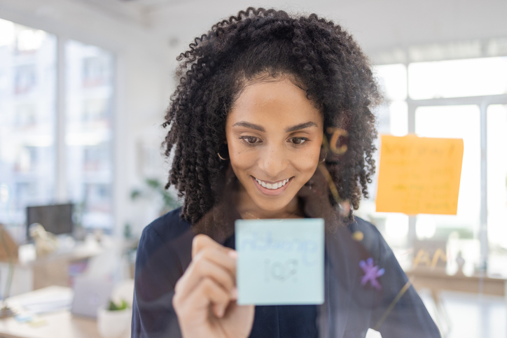 Planning, thinking and writing by black woman in office for agenda, calendar or schedule on sticky note. Creative, strategy or lady with idea for future, vision or innovation, goal or problem solving