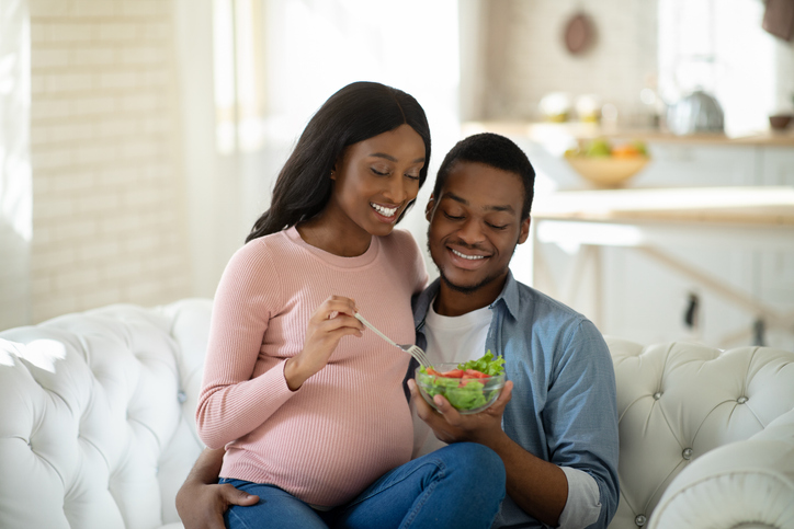 Healthy nutrition during pregnancy. Positive black husband and his expectant wife eating fresh vegetable salad at home