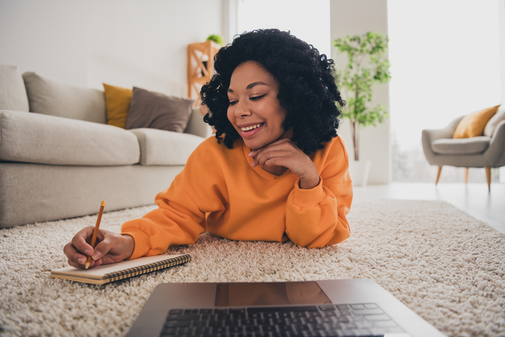Photo of lovely young lady lying floor study remotely write notes dressed casual orange clothes cozy day light home interior living room