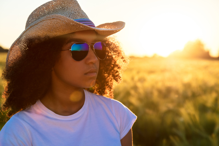 Beautiful happy mixed race African American female girl teenager young woman wearing relective aviator sunglasses and cowboy hat in a cornfield at golden sunset or sunrise