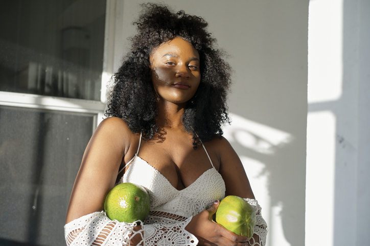 Woman in white crochet top holding two large green fruits, standing in sunlight. Miami, Florida, USA