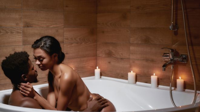 Romantic evening. Young sensual naked african couple of lovers kissing while taking foamy bath with candles together