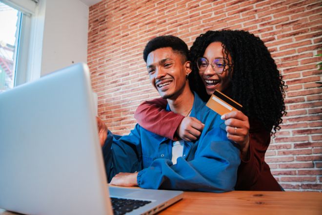 A cheerful African American couple is using a laptop to search a sale on a online shop. Excited husband and wife are sitting in the living room, with a credit card looking on a computer for a delivery
