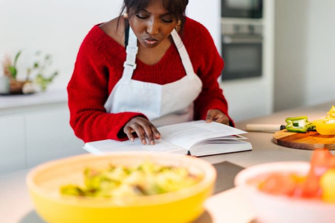 Woman wearing apron reading cookbook and preparing vegetables in kitchen
