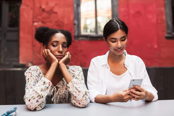Upset african girl sadly looking aside while asian girl near happily using cellphone in courtyard of cafe