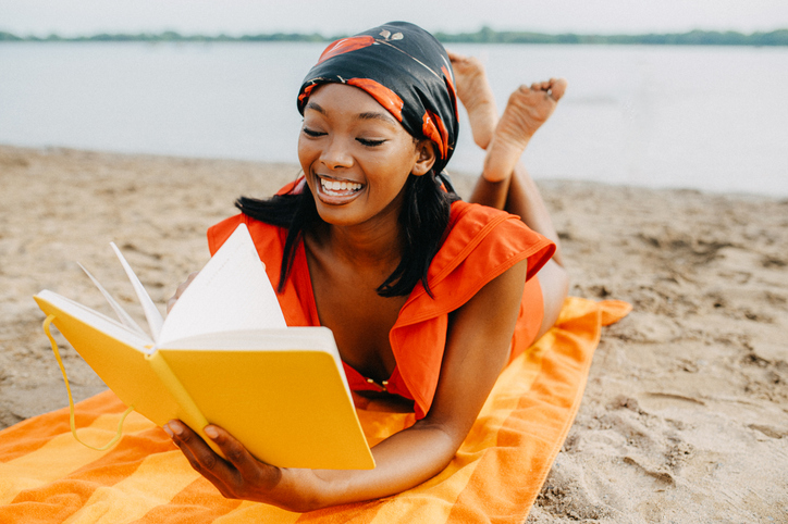 A woman lies on an orange towel at the beach, reading a book with a bright yellow cover.