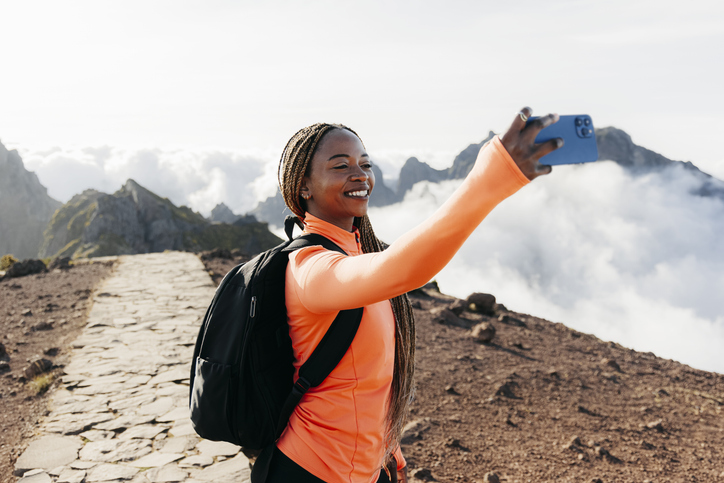 Young woman with a backpack and long braids taking a selfie with her cell phone on a hiking trail